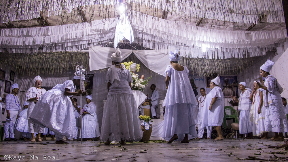 Imagem colorida de um ritual em um terreiro de candomblé. Em um salão espaçoso, vários homens e mulheres vestindo roupas brancas se reúnem e interagem em forma de círculo. No teto, várias fitas de cor branca e, no chão, várias folhas secas espalhadas.