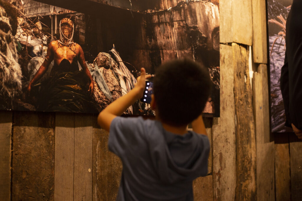 Em um espaço interno com paredes de tábuas, estão expostas fotografias grandes com performers vestidos como entidades afro-indigenas. Diante deles, uma criança tira uma foto com uma câmera digital.