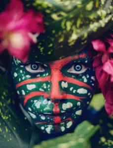 Imagem colorida de Uýra Sodoma com maquiagem verde com pontos brancos e traços vermelhos. Ela está no meio de folhas e flores
