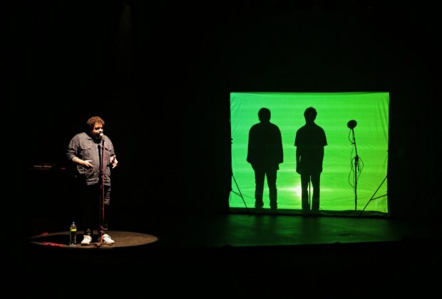 Imagem colorida de um palco escuro. Do lado esquerdo, um homem vestindo um macacão diante de um microfone. Do lado esquerdo, as silhuetas de dois homens aparecem diante de uma tela com uma projeção verde.