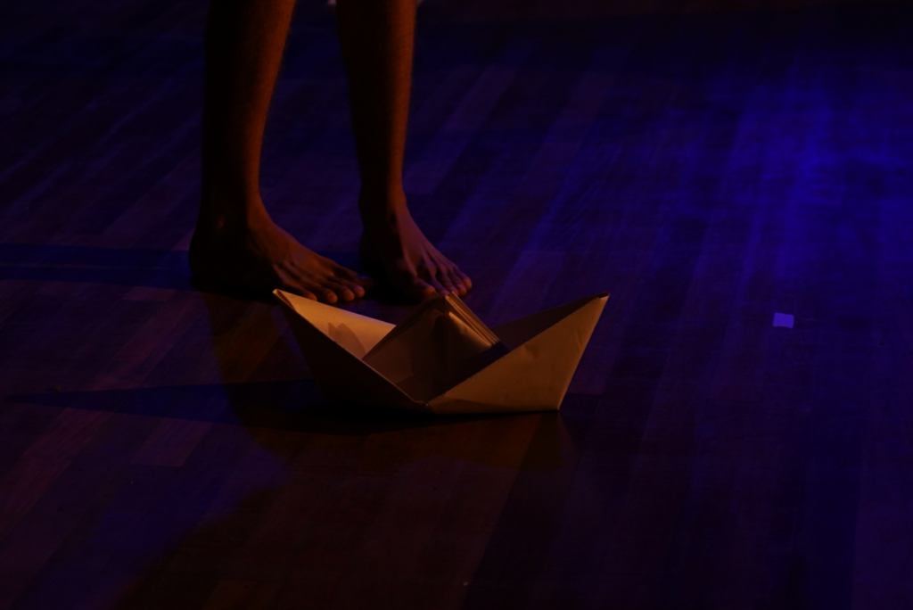 Imagem colorida do chão de um palco. Nele, um par de pés de um jovem. Diante deles, um barco de papel.