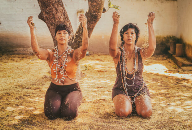 Imagem colorida de um quintal com chão de terra. Nele, estão duas mulheres com feições indígenas, Iris e Iara Campos. Elas estão ajoelhadas e erguem os dois braços ao lado do corpo.