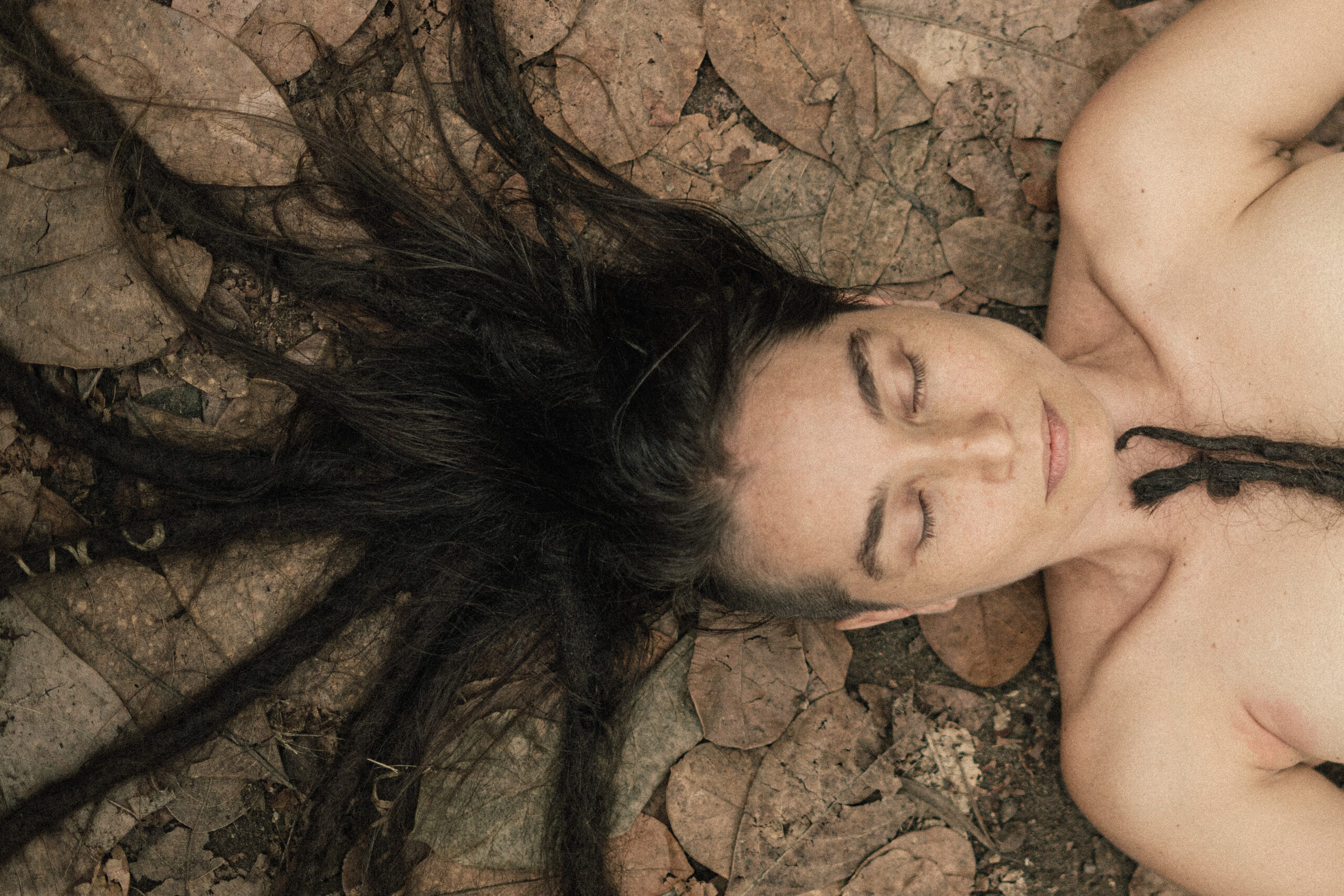 Imagem colorida de mulher deita em um chão rachado. Ela está com o torso despido e os cabelos espalhados pelo chão.