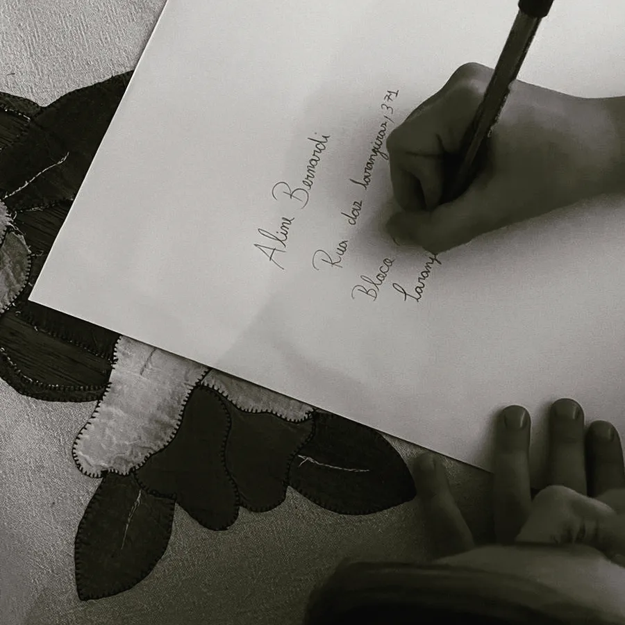 Imagem em preto e branco de uma carta sendo escrita por uma mão. No papel, está escrito do endereço da autora Aline Bernardi.