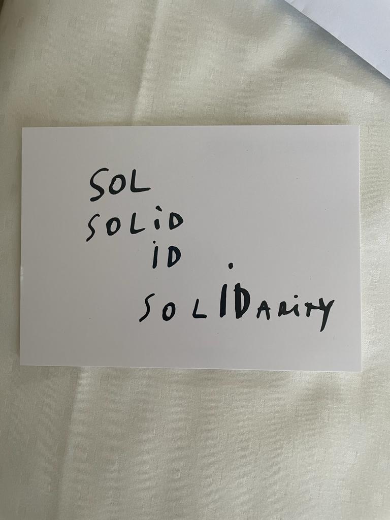 Imagem colorida de um postal na cor branca. Nele, está escrito uma espécie de poema concreto: um arranjo entre as palavras 'Sol', 'Solid', 'Id' e 'Solidary'.