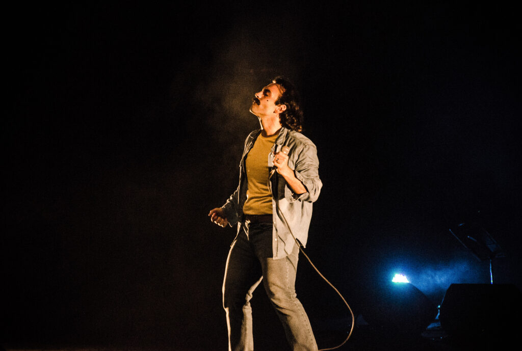 Imagem colorida de um palco escuro. No centro, um homem branco com cabelo e bigode negros. Ele usa calça jeans, jaqueta e uma camisa amarelada e segura um microfone na mao esquerda.