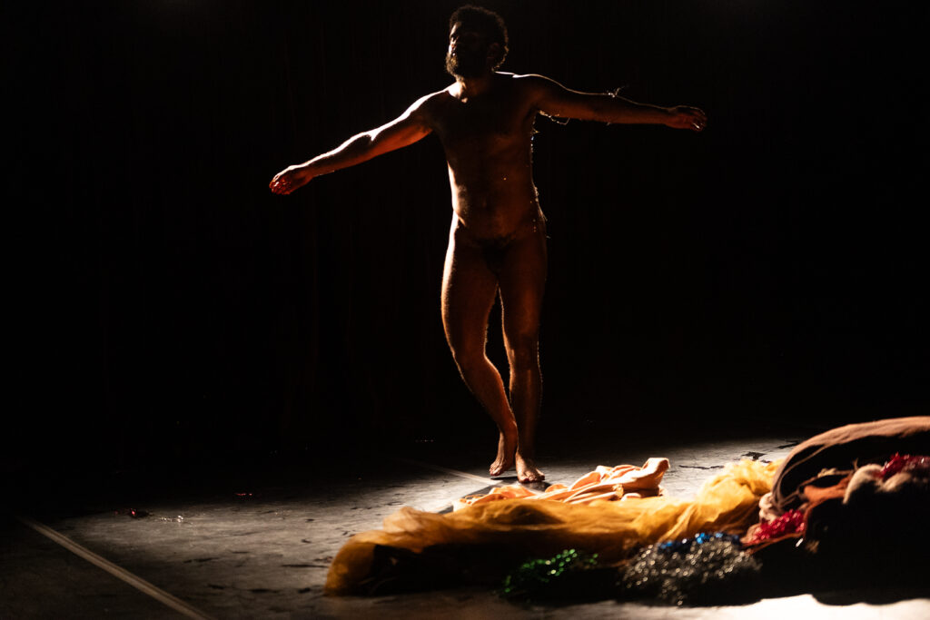 Imagem colorida de um palco escuro. Nas sombras, um homem negro seminu, usando uma tanga, está com os braços abertos. No chão à direita, está um amontoado de tecidos com texturas distintas.