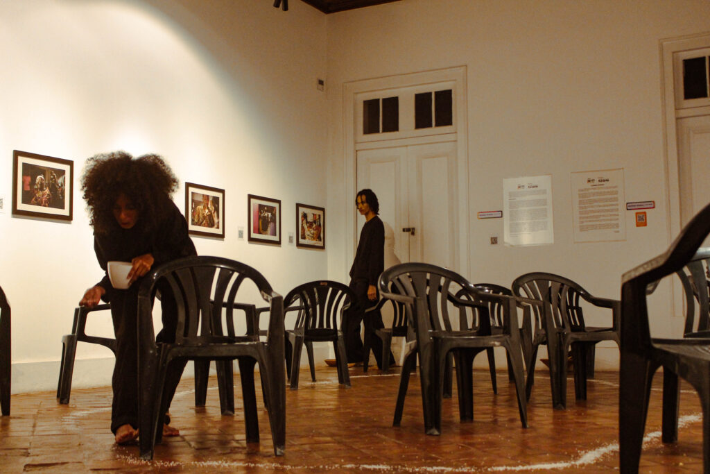 Imagem colorida de uma sala com chão de taco e paredes brancas e alguns quadros pequenos pendurados. O espaço é repleto de cadeiras pretas e, dentre elas, caminham um homem e uma mulher negros. A mulher aparece em primeiro plano, derramando sal no chão. O homem aparece ao fundo da imagem, observando a mulher.