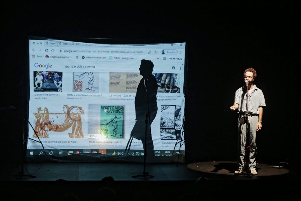 Cena de um espetáculo teatral com duas figuras no palco. À direita, um ator vestido com macacão cinza, segurando um microfone, gesticula enquanto fala. Ao fundo, há uma grande projeção com uma página do Google aberta, exibindo resultados de pesquisa para "Morte e Vida Severina", incluindo capas de livros e ilustrações relacionadas ao poema de João Cabral de Melo Neto. À esquerda, a silhueta de outra pessoa, em frente à projeção, contrasta com a luz projetada. O ambiente é escuro, com iluminação focada no ator e na projeção.