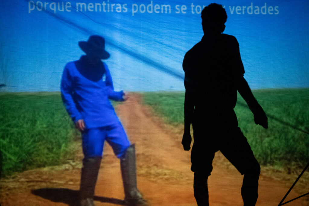 Cena de um espetáculo teatral com uma projeção ao fundo que mostra um homem vestido com macacão azul, chapéu preto e botas, em uma estrada de terra no meio de um campo de plantação. Acima da imagem, aparece a frase: "porque mentiras podem se tornar verdades". Em primeiro plano, à direita, a silhueta de um performer é visível, de costas ou de perfil, criando um contraste com a iluminação da projeção. A interação entre a sombra do ator e a projeção cria uma composição visual dinâmica.