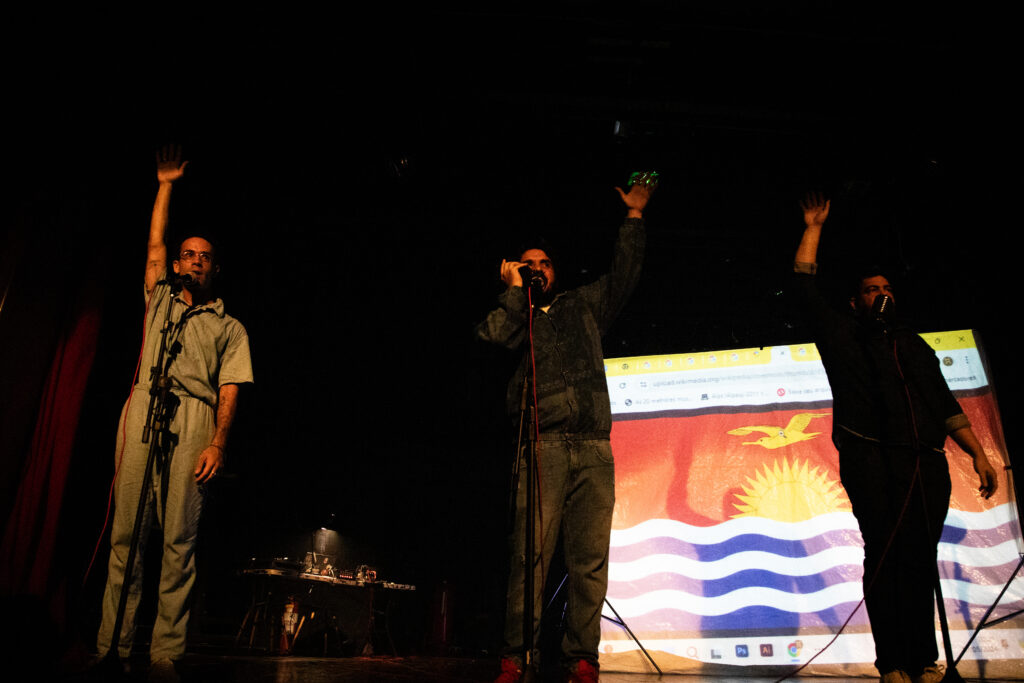 Três atores em um palco, cada um com um microfone e com um dos braços levantados. À esquerda, um performer veste um macacão cinza; ao centro, outro com roupas casuais; e à direita, o terceiro performer usa uma camisa escura. Ao fundo, uma projeção exibe a bandeira de Pernambuco parcialmente visível, com o céu azul, o sol dourado e ondas brancas e azuis. Também é possível identificar detalhes de uma interface de navegador web acima da bandeira. 